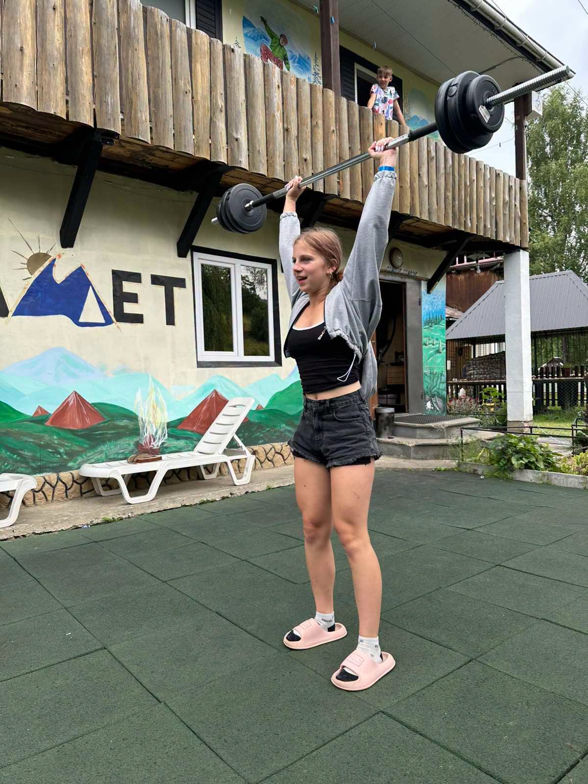 Дівчина виконує жим штанги над головою на спортивному майданчику табору.