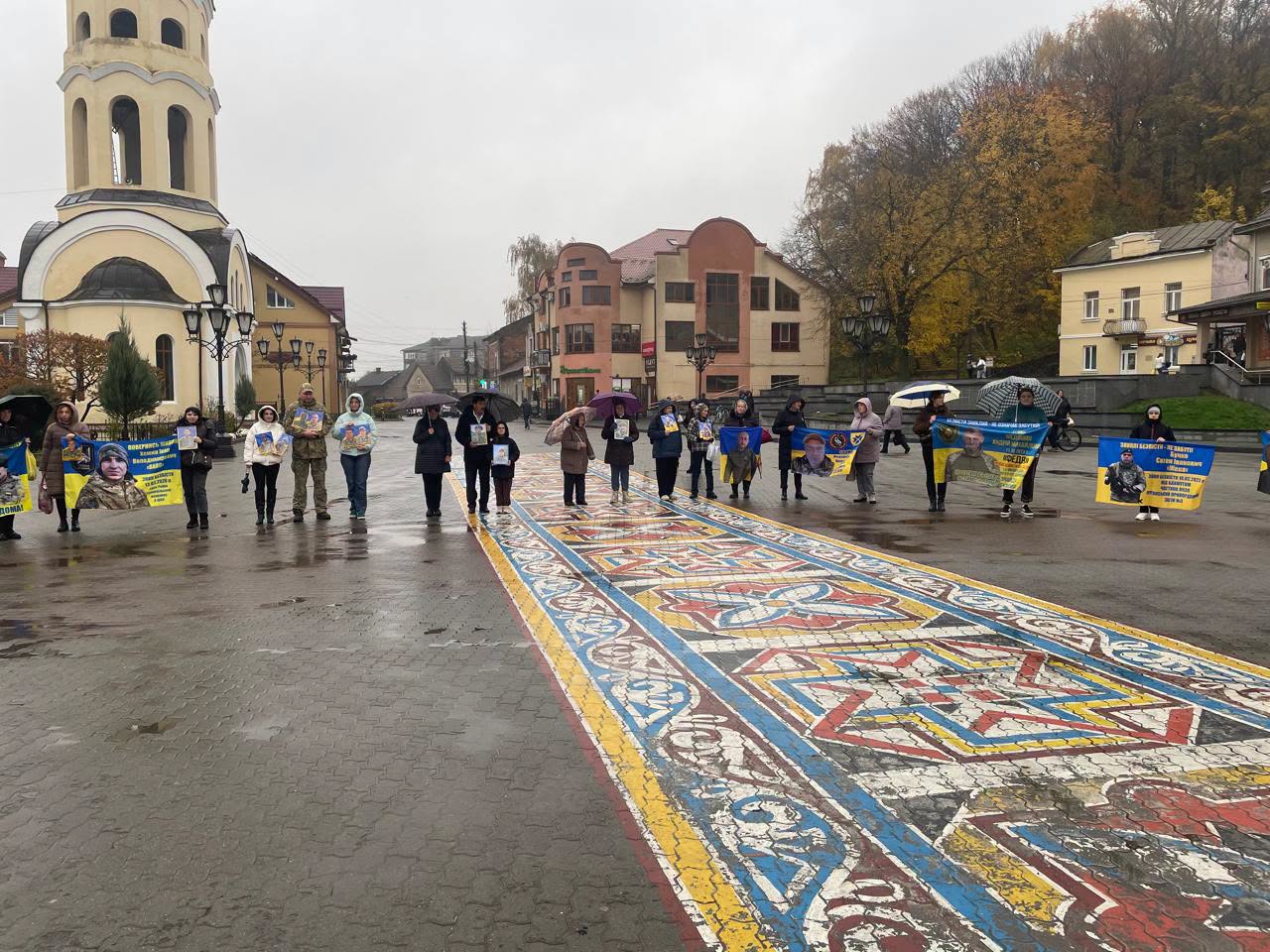 Люди з портретами військових і синьо-жовтими плакатами стоять на площі